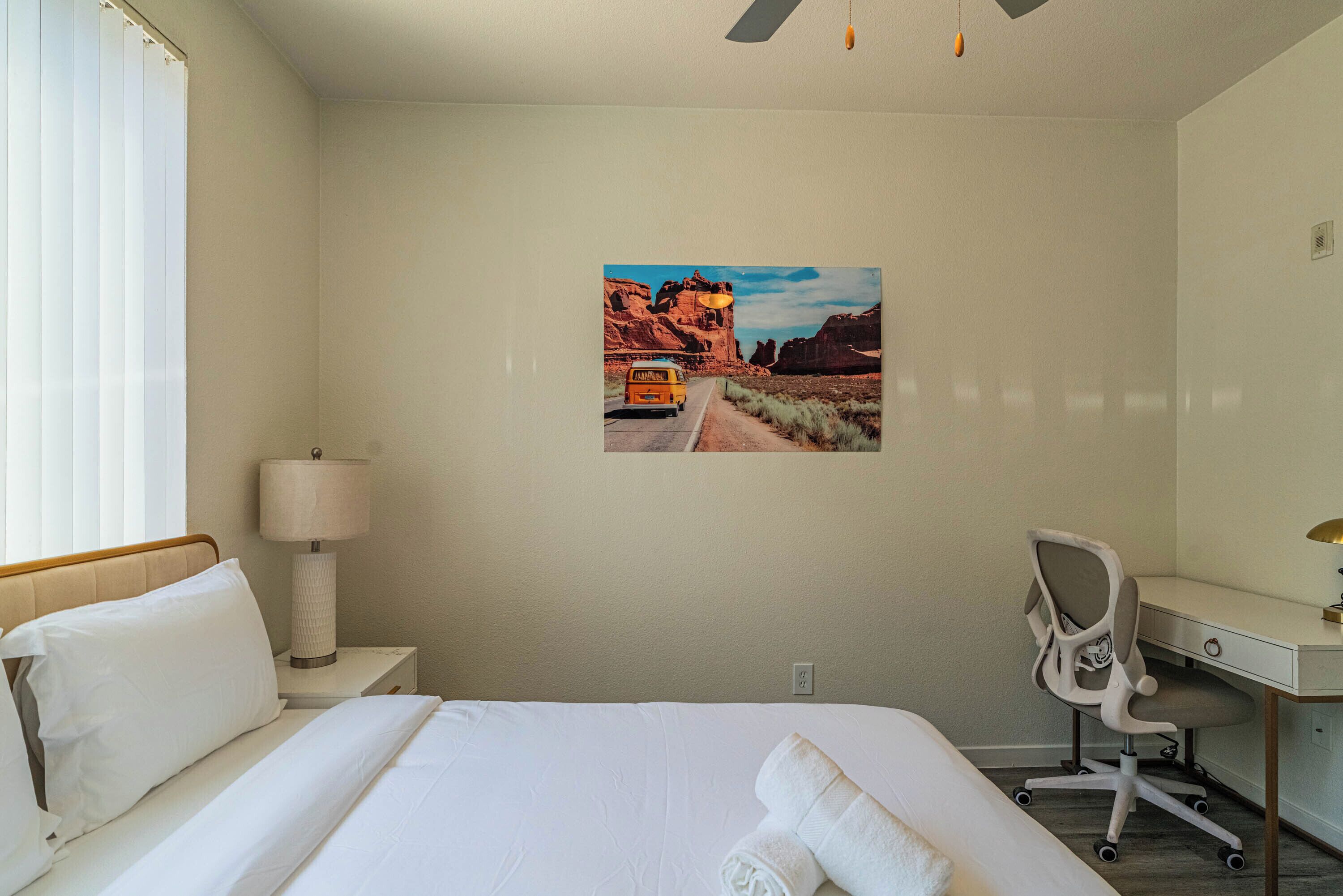 A hotel room with a bed, a desk, a chair, a lamp, and a painting of a desert road.
