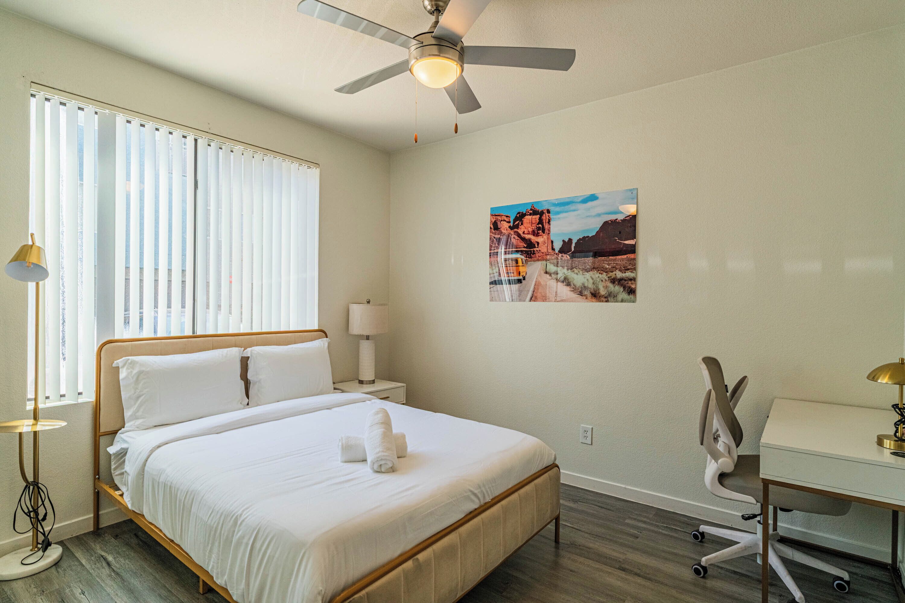 Une chambre d’hôtel comprenant un lit, un bureau, une chaise, un ventilateur de plafond, une fenêtre avec des stores et un tableau accroché au mur.