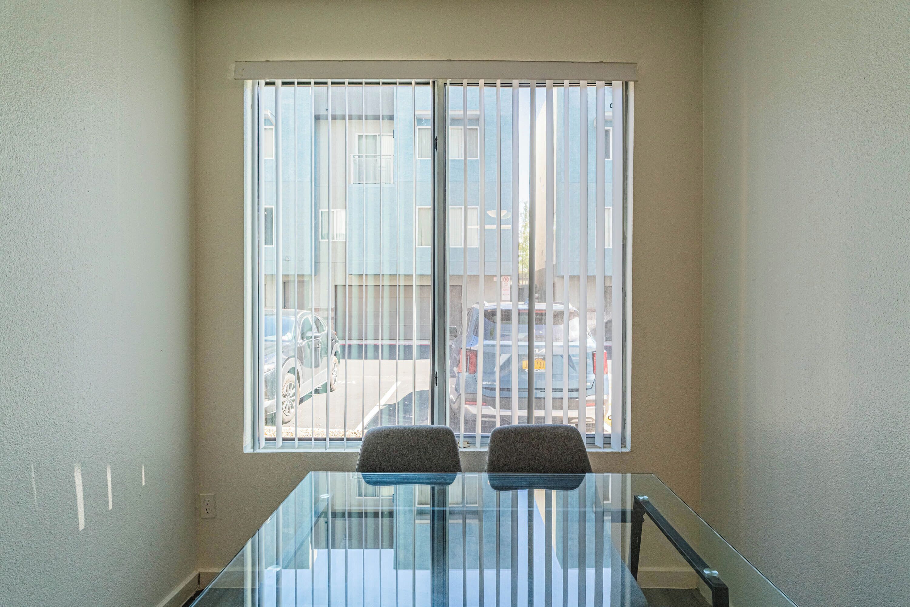 A room with a glass table and two chairs, a window with blinds, and a view of parked cars outside.