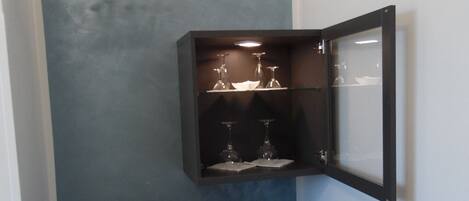 A compact room with a built-in wardrobe, a small cabinet with wine glasses, and a refrigerator.