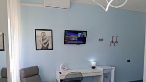 Une pièce avec un canapé gris, un bureau avec un ordinateur et une lampe, un téléviseur fixé au mur et un tableau encadré.