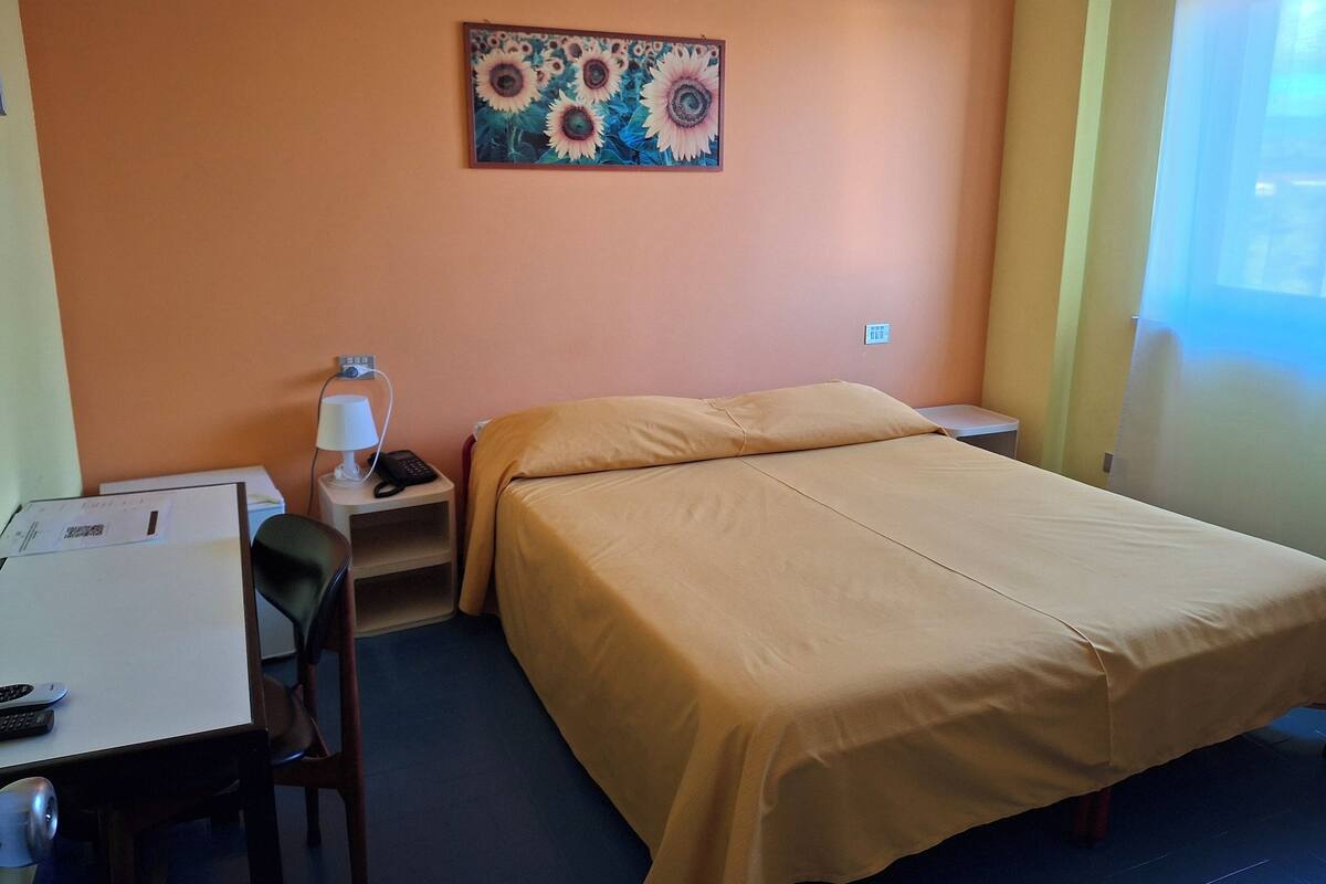 A hotel room with a bed, a desk with a chair, a lamp, and a painting on the wall.