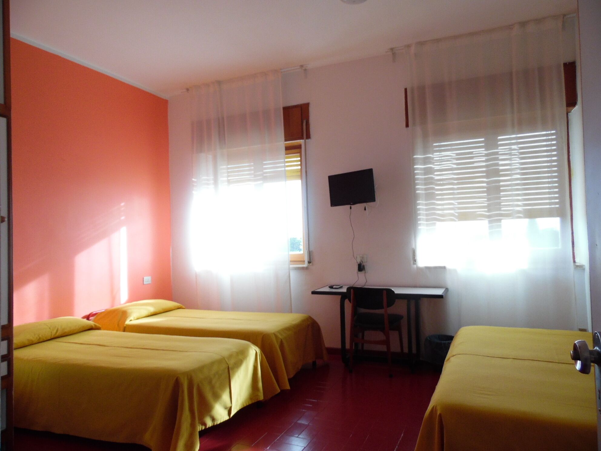A room with two single beds, a desk with a chair, a television mounted on the wall, and a window with blinds.