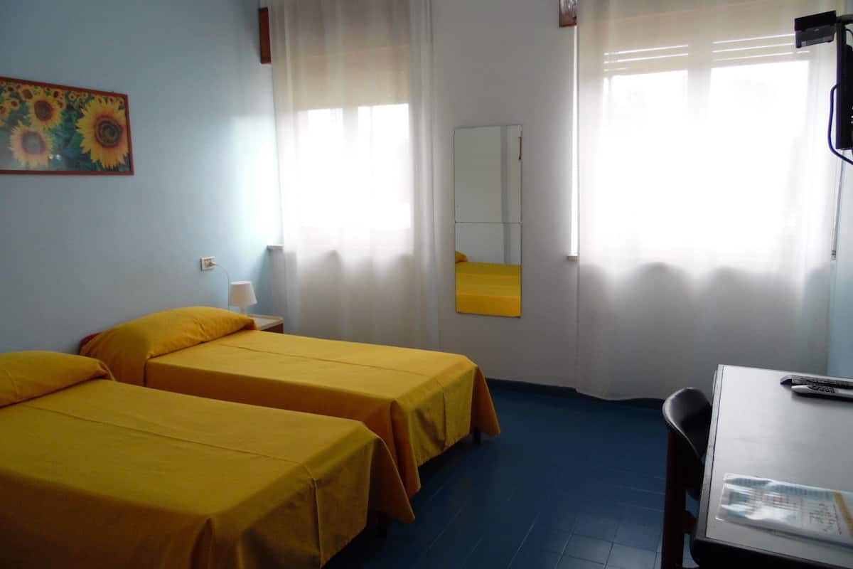 A room with two beds, a desk, a mirror, and a window with sheer curtains.