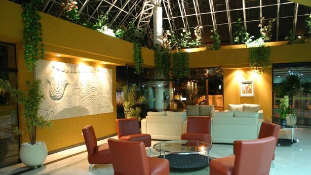 Un hall d’entrée spacieux avec un plafond haut, comprenant un espace salon central avec des fauteuils rouges et une table basse en verre. Les murs sont ornés d’une grande fresque décorative et de plantes en pot.