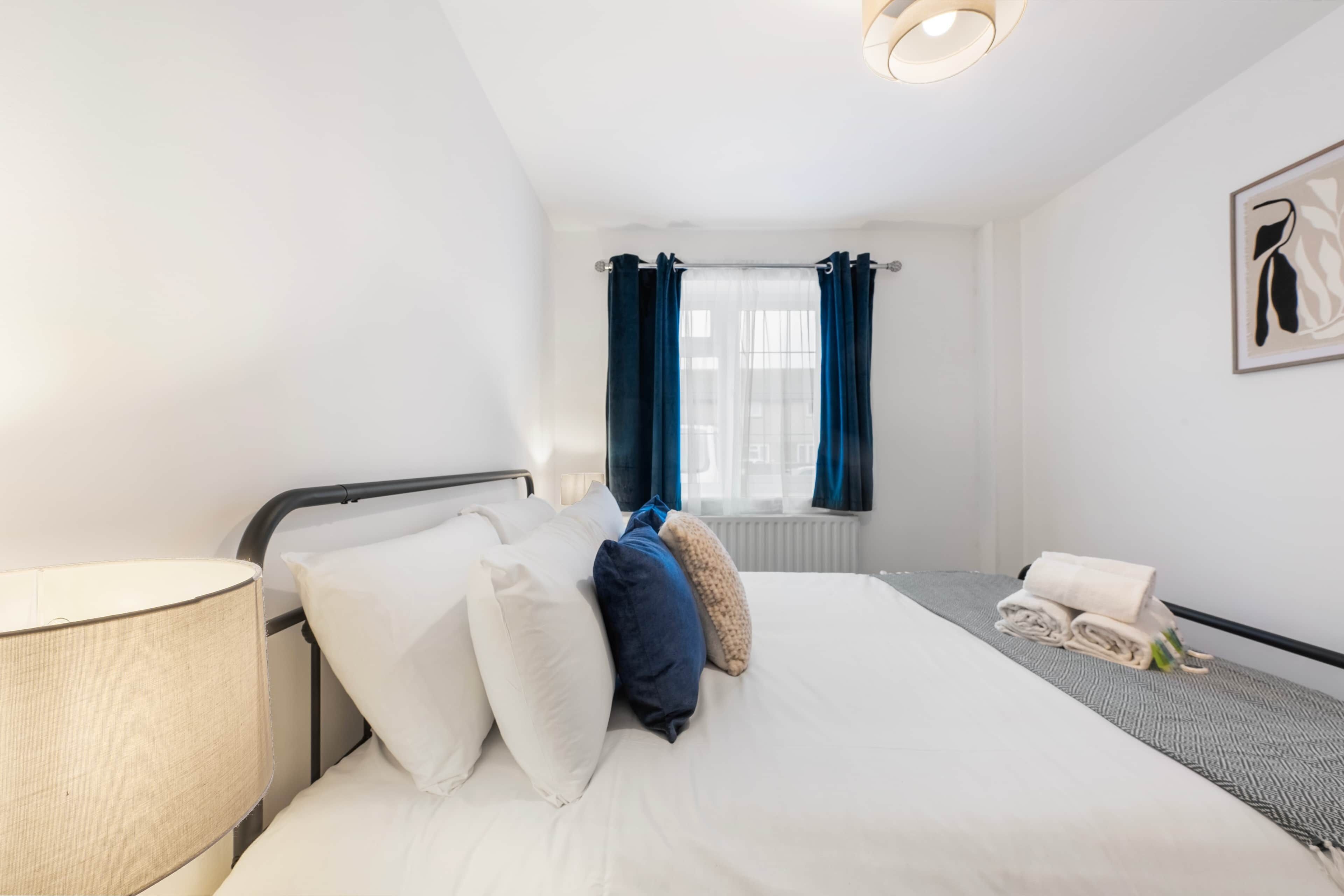 Een net opgemaakt bed met witte en blauwe kussens, een bedlamp en een opgerolde handdoek op een grijze loper.