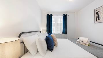 Een net opgemaakt bed met witte en blauwe kussens, een bedlamp en een opgerolde handdoek op een grijze loper.