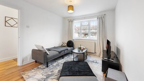 Ein modernes, helles Zimmer mit einem grauen Sofa, einem kleinen runden Tisch, einem schwarzen Ottomanen und einem Fernseher auf einem schwarzen Schrank.