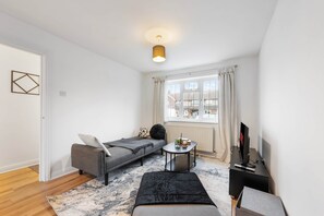A modern, well-lit room with a grey sofa, a small round table, a black ottoman, and a TV on a black stand. - Urban Stay (Slough)