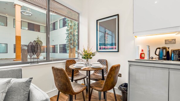 A modern dining area with a round table, cushioned chairs, and a view of the building outside. - The Cosy Luxe (Slough)