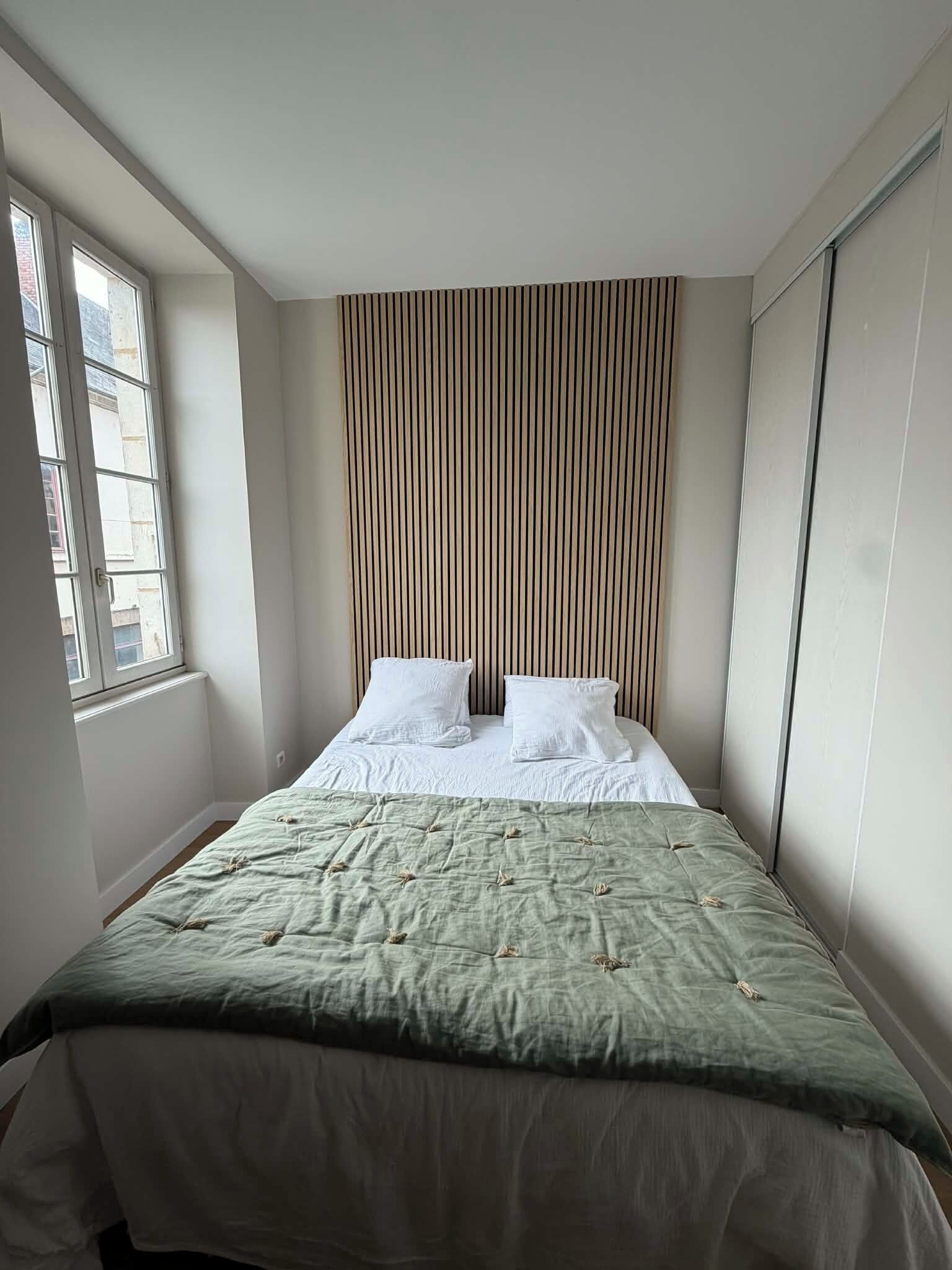 Un lit bien fait, avec une literie blanche et un plaid vert, une fenêtre sur la gauche et un panneau de bois vertical sur le mur.