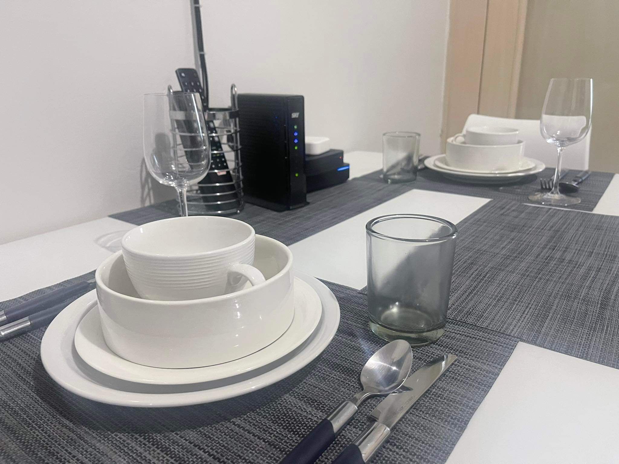 A neatly set dining table with white plates, bowls, and cutlery, a glass of water, and a remote control on the table.