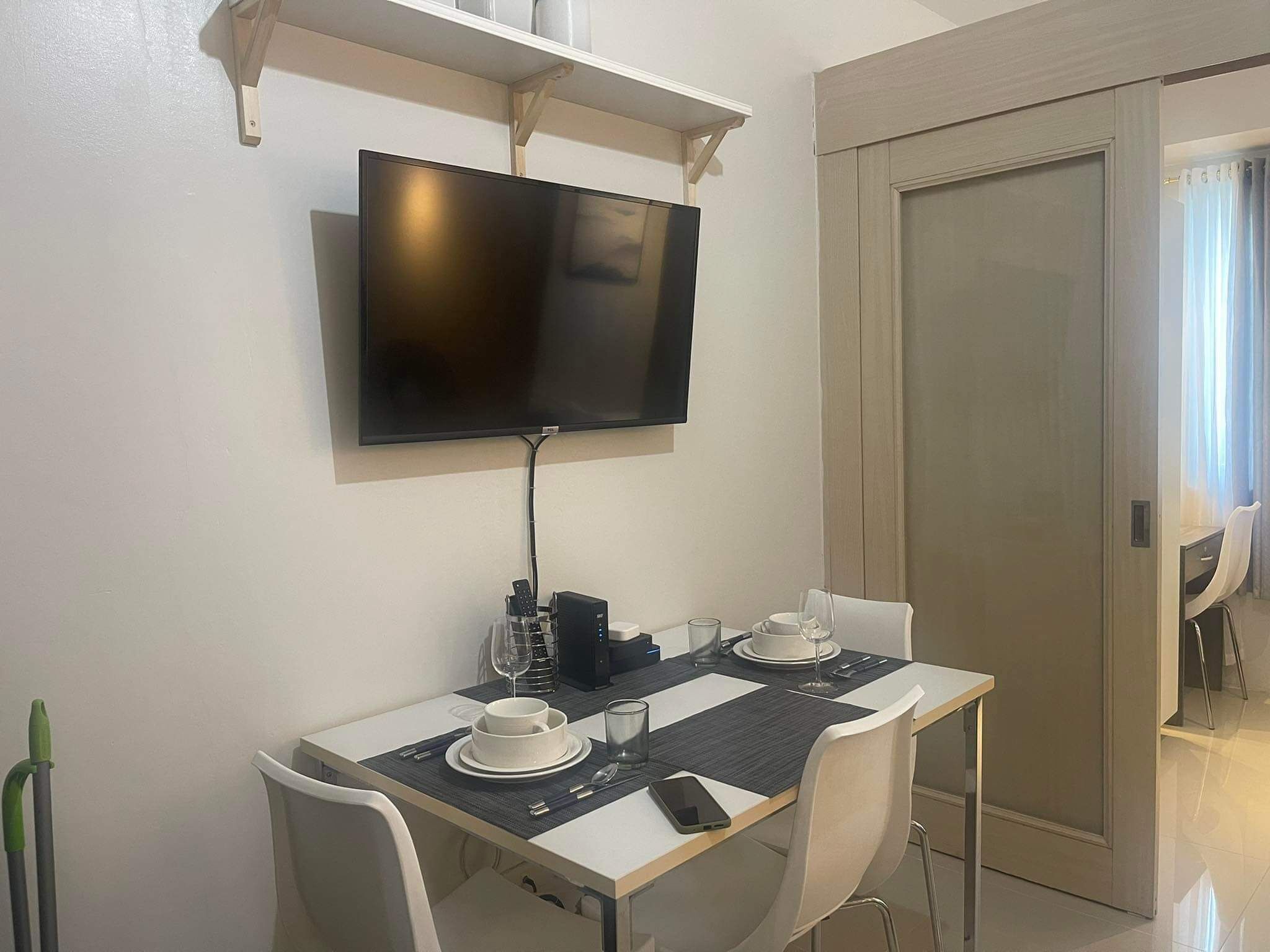 A dining area with a table set for two, a television mounted above the table, and a door to the right.