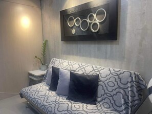 Ein modernes Schlafzimmer mit einem grauen gemusterten Sofa, zwei dunklen Kissen, einem kleinen Nachttisch mit Pflanze und einem an der Wand befestigten abstrakten Kunstwerk.