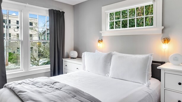 A bedroom with a large bed, a window with curtains, a bedside table with a lamp, and a wall-mounted light fixture. - Montlake Hideaway in Spite House (Seattle)