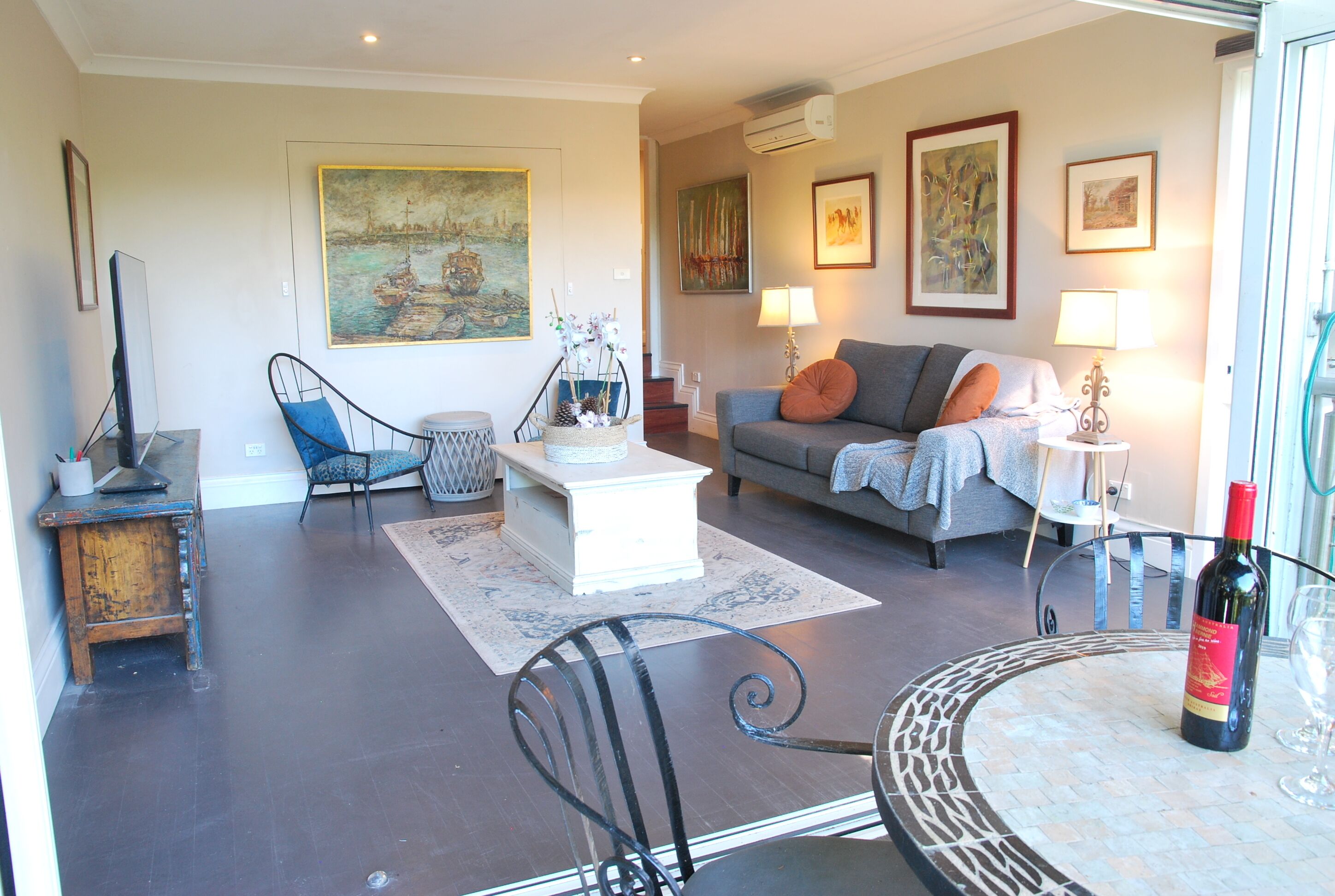 Un salon comprenant un canapé, des fauteuils, une table basse et une table à manger avec des chaises. Les murs sont ornés de tableaux, il y a une télévision et une bouteille de vin sur la table.
