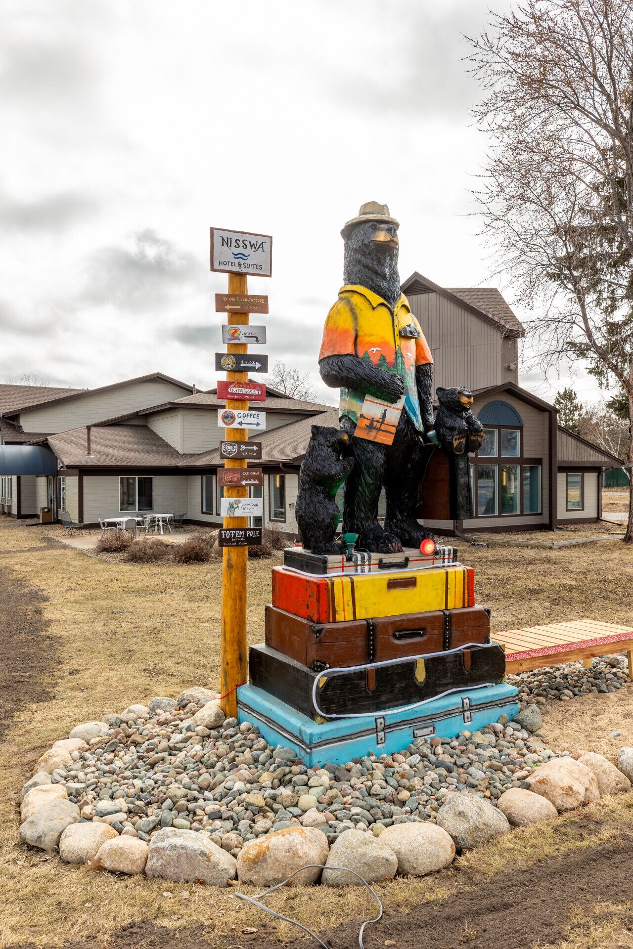 Une statue d’ours avec des valises, un panneau indicateur et un bâtiment en arrière-plan.