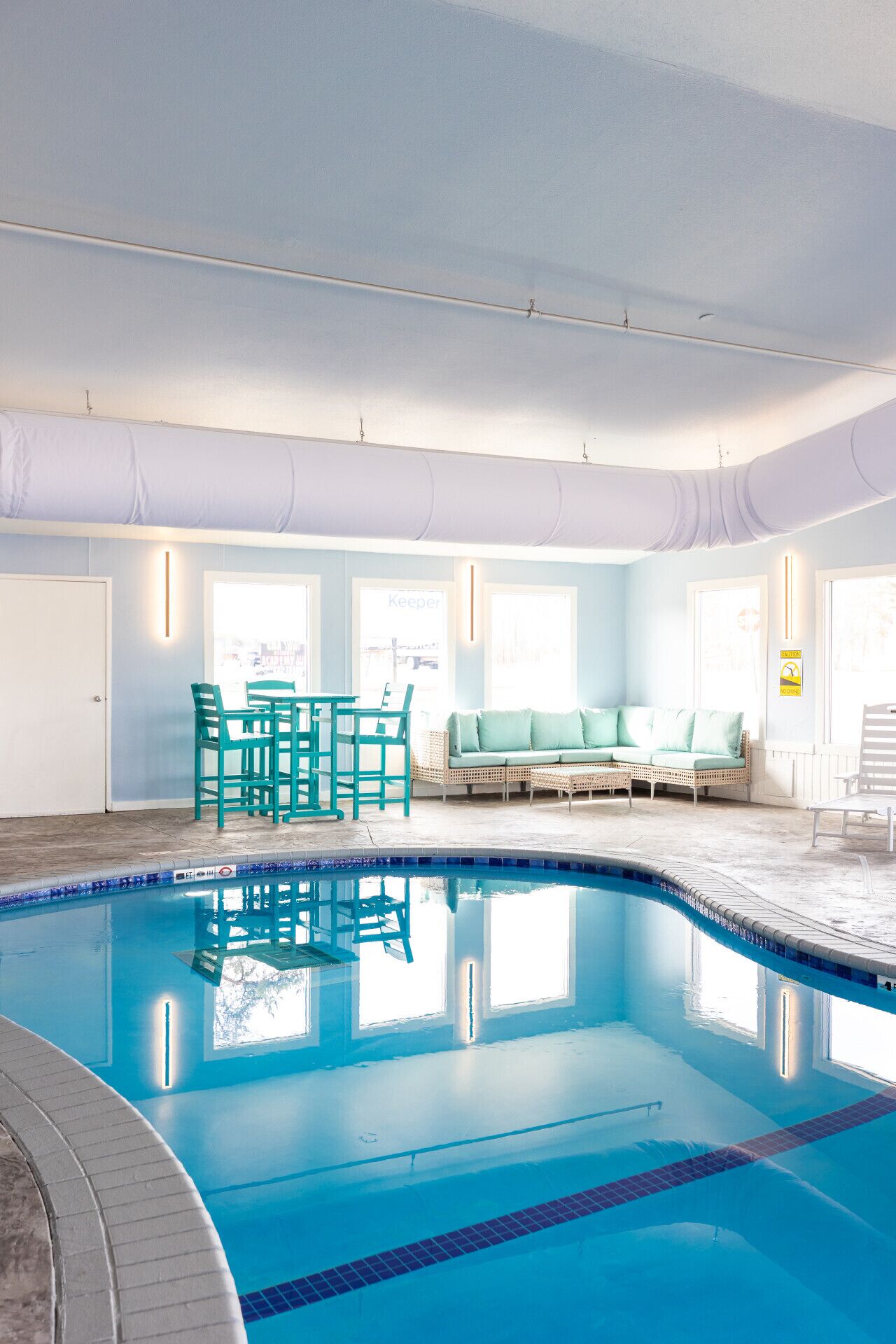 Una piscina interior con agua azul cristalina, una zona de estar con sofá y sillones, y grandes ventanales que dejan entrar luz natural.