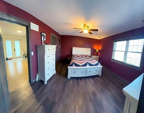 A bedroom with a bed, dresser, ceiling fan, window, and a door leading to another room.