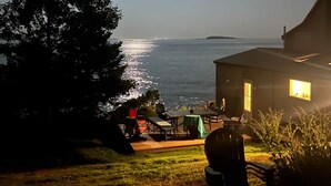 A house with a deck overlooking a body of water, a tree to the left, and a person sitting on a chair. - Barnacle - quiet family Maine waterfront getaway (Harpswell)