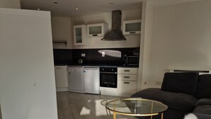 A modern kitchen with white cabinets, a black countertop, and a built-in oven. There is a glass coffee table with gold legs in the living area.