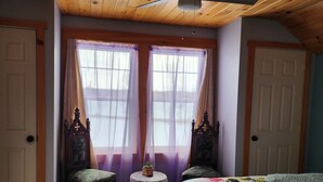 A bedroom with wooden ceiling, two windows with sheer curtains, a bed with a floral bedspread, a small round table with a basket, and two chairs. - Lakeside oasis Apt with water access and views.
There is a She shed on the beach (Norwood)