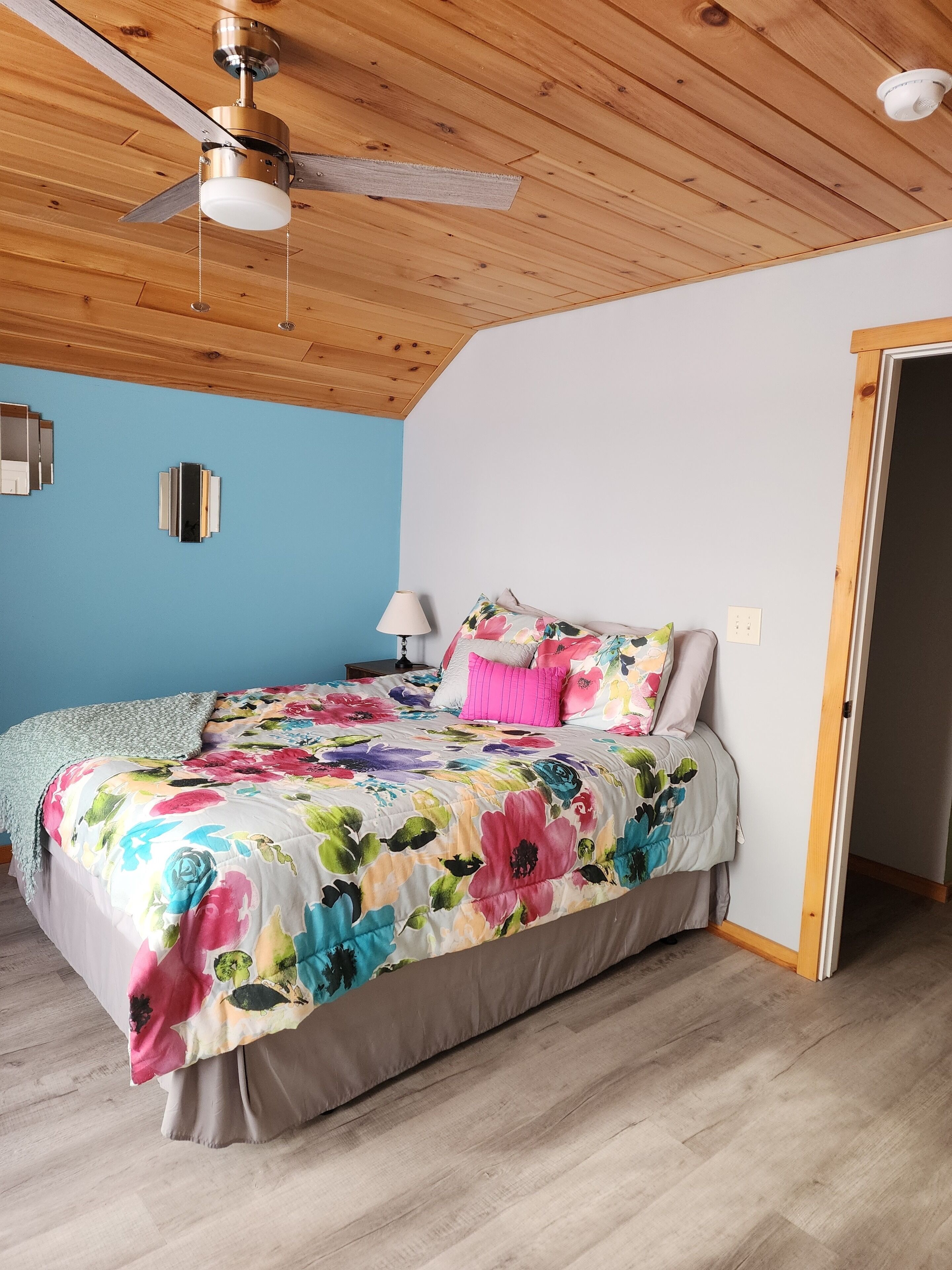 Une chambre à coucher avec un plafond en bois, un lit recouvert d’une couvre-lit à motifs floraux, une table de chevet et une lampe.