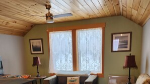 A cozy living room with a wooden ceiling, a large window with lace curtains, a sectional sofa, a patterned armchair, and a dining table. - Lakeside oasis Apt with water access and views.
There is a She shed on the beach (Norwood)