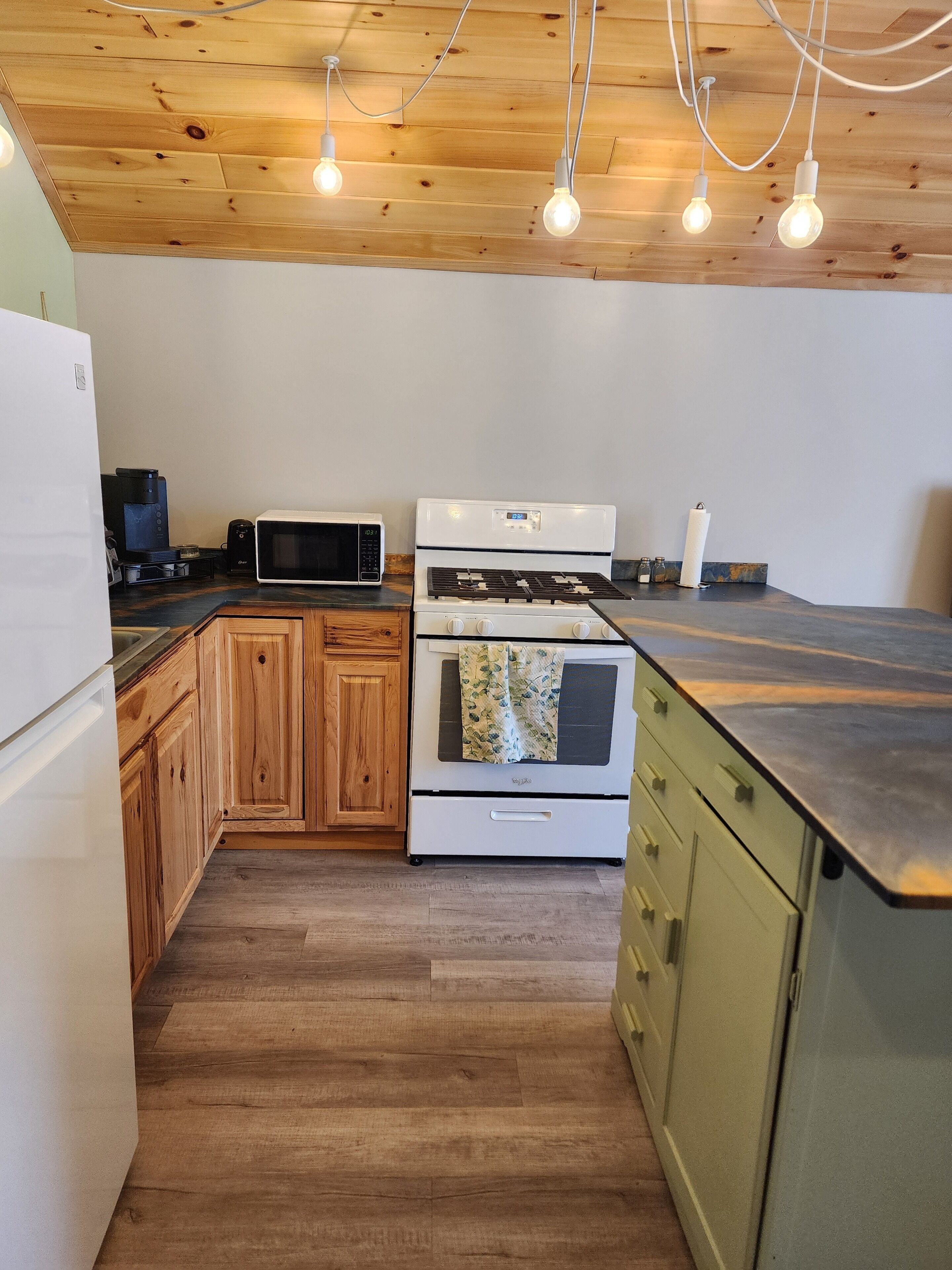 A kitchen with wooden cabinets, a white refrigerator, a microwave, a stove, and hanging light bulbs.
