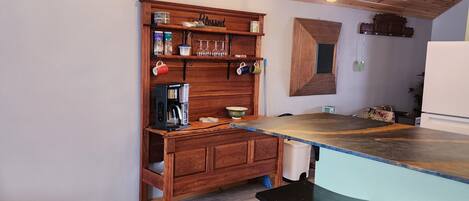 A kitchen with wooden ceiling, a wooden shelf with various items, a coffee maker, and a bar counter with stools.