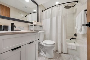 Une salle de bain moderne avec des toilettes blanches, un lavabo doté d’un robinet noir, une douche avec un rideau blanc et une baignoire avec une serviette blanche.
