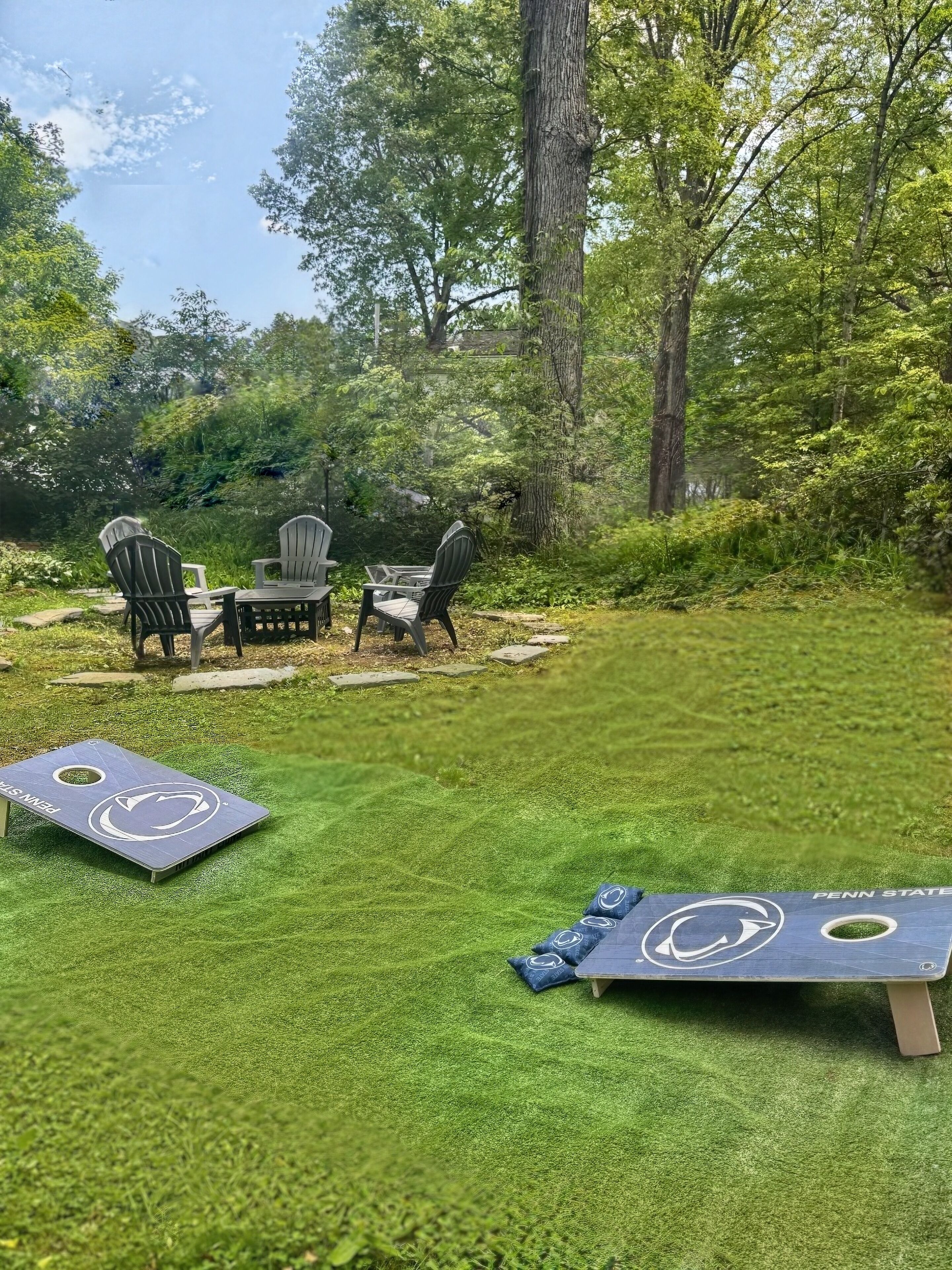 An outdoor seating area with Adirondack chairs and a table, surrounded by a lush green lawn and trees.