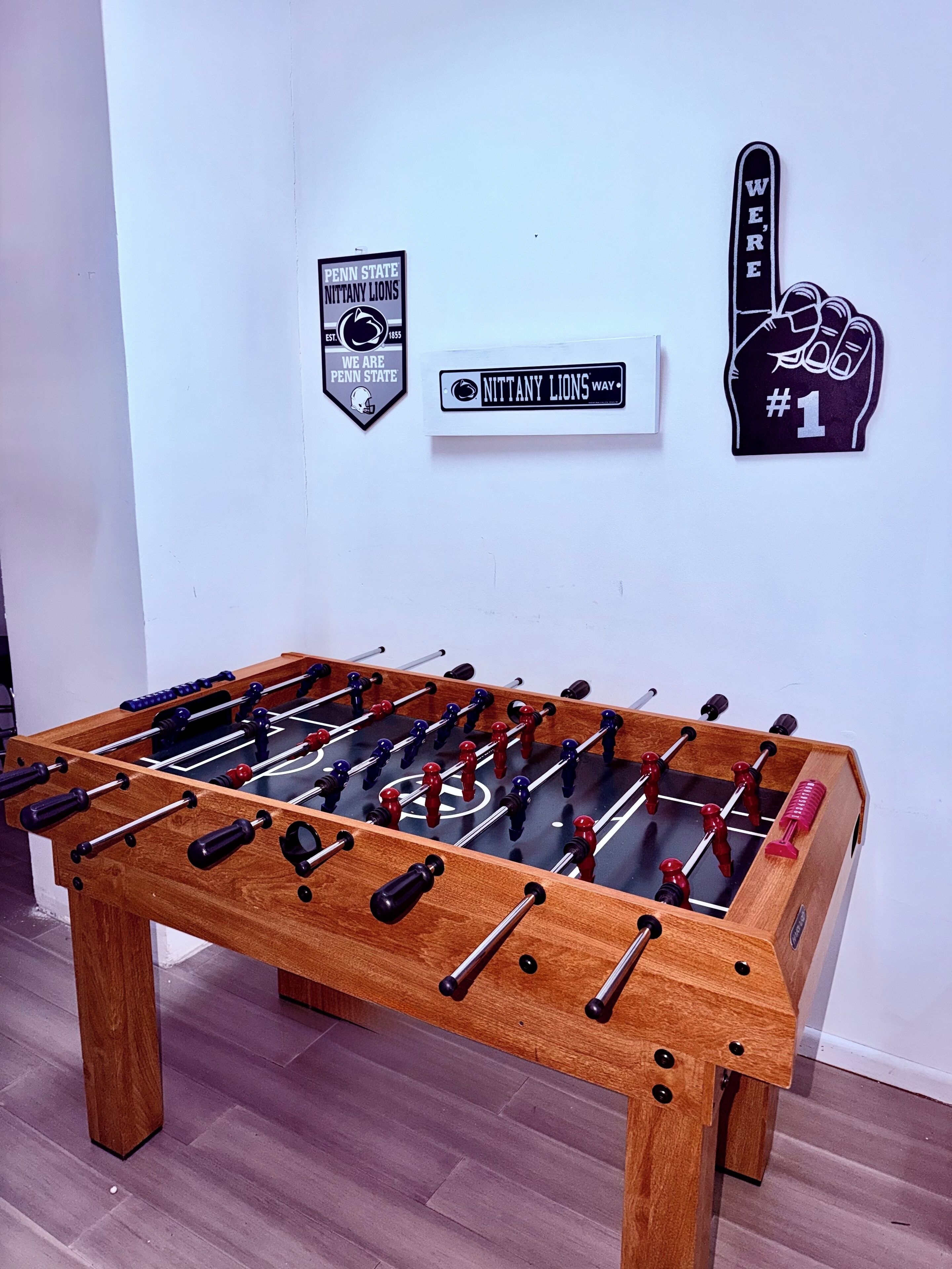 Een tafelvoetbaltafel in een kamer met een witte muur. Op de muur hangen drie borden: één met 'Penn State Nittany Lions', een ander met 'Nittany Lions Way' en een derde met 'We're Penn State'.