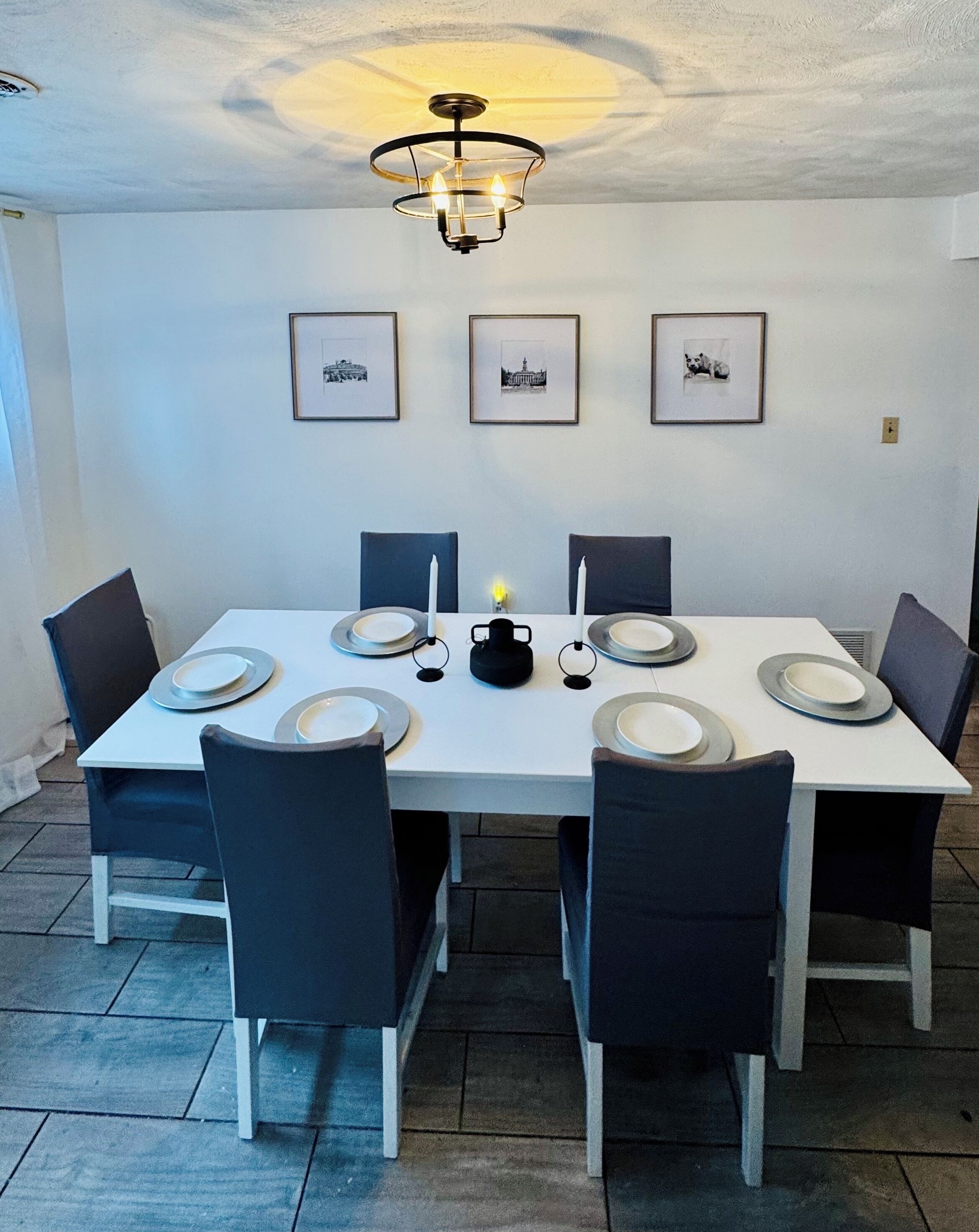 Un coin repas avec une table blanche dressée pour six personnes, des chaises noires et un lustre. Trois tableaux encadrés sont accrochés au mur au-dessus de la table.