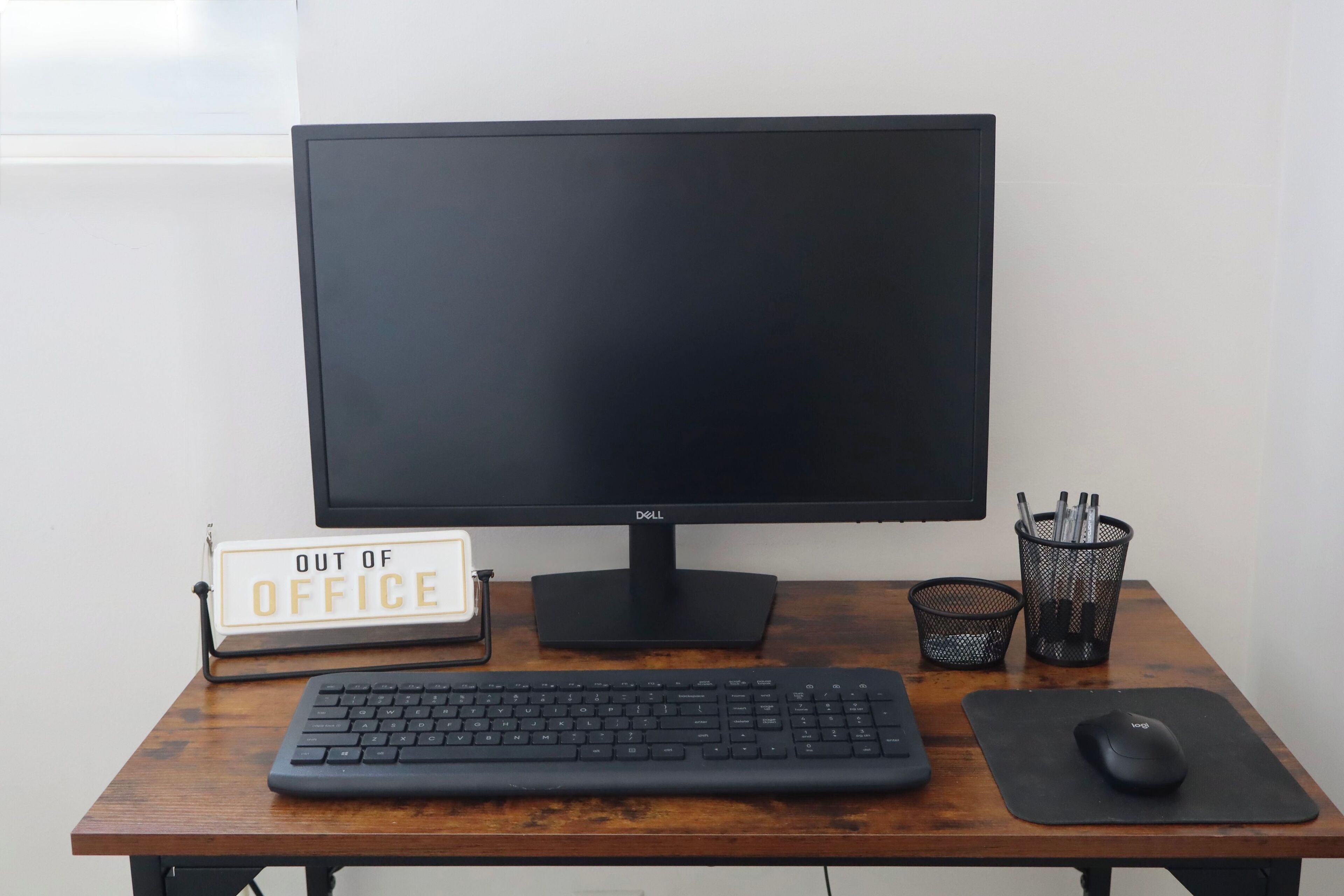 Un bureau en bois avec un écran Dell noir, un clavier noir, une souris, un panneau indiquant « ABSENT » et un porte-stylos.