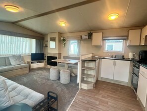 Interior compacto de um veículo recreativo, com sofá, televisão, pequeno espaço de cozinha e área de jantar.