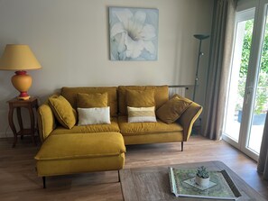 Een mosterdgele bank met meerdere kussens, een klein houten bijzettafeltje met een lamp, een lijst met bloemkunst aan de muur, en een potplant op een houten dienblad.