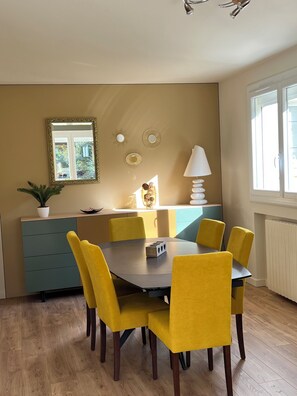 Een eethoek met een ronde tafel en zes gele stoelen, een blauwe kast, een potplant en een raam met uitzicht op groen.