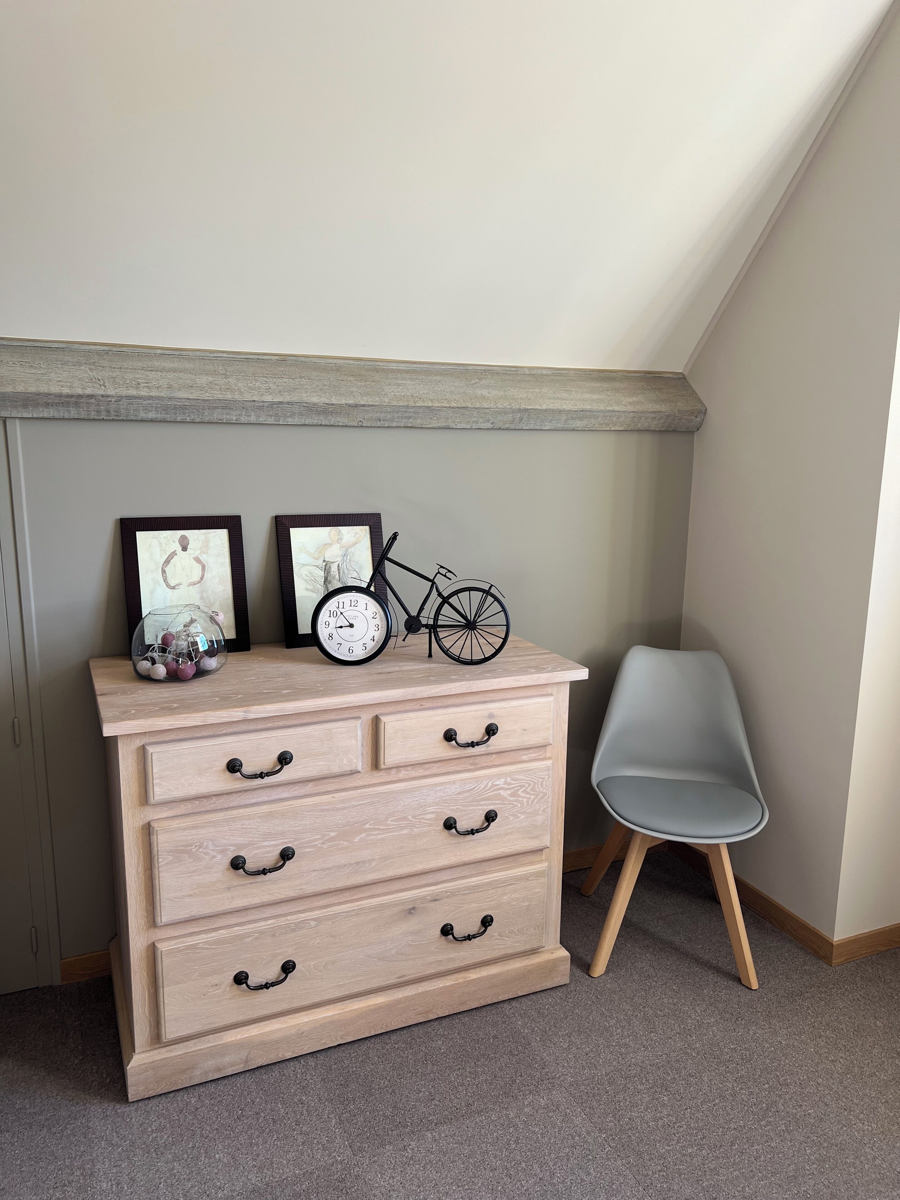 A corner room with a wooden dresser, two framed pictures, a bicycle, and a grey chair.