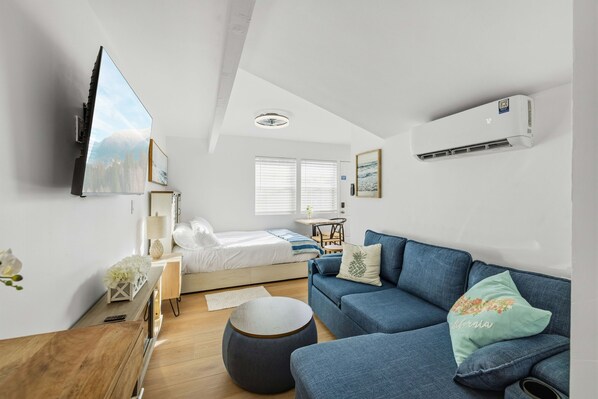 A modern, well-lit room with a bed, a blue sofa, a wooden table, and a mounted TV.