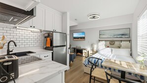 Eine moderne Küche mit weißen Schränken, einem Edelstahl-Kühlschrank und einer Marmorarbeitsplatte. Es gibt einen Essbereich mit einem Holztisch und Stühlen, sowie ein Bett mit einer blau-weiß gestreiften Decke.