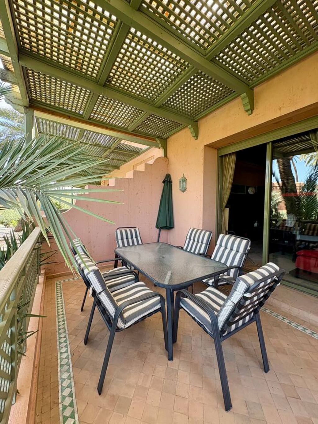An outdoor patio with a table and chairs, a pergola overhead, and a palm tree.