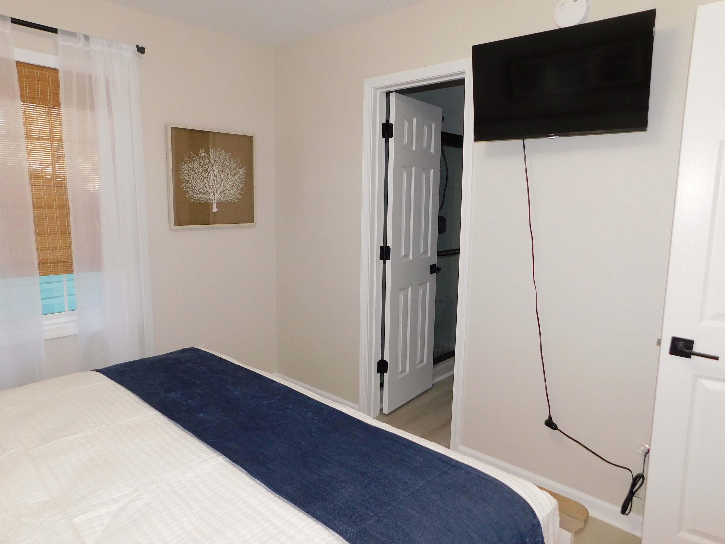 A bedroom with a bed, a TV mounted on the wall, a door leading to another room, and a window with blinds.