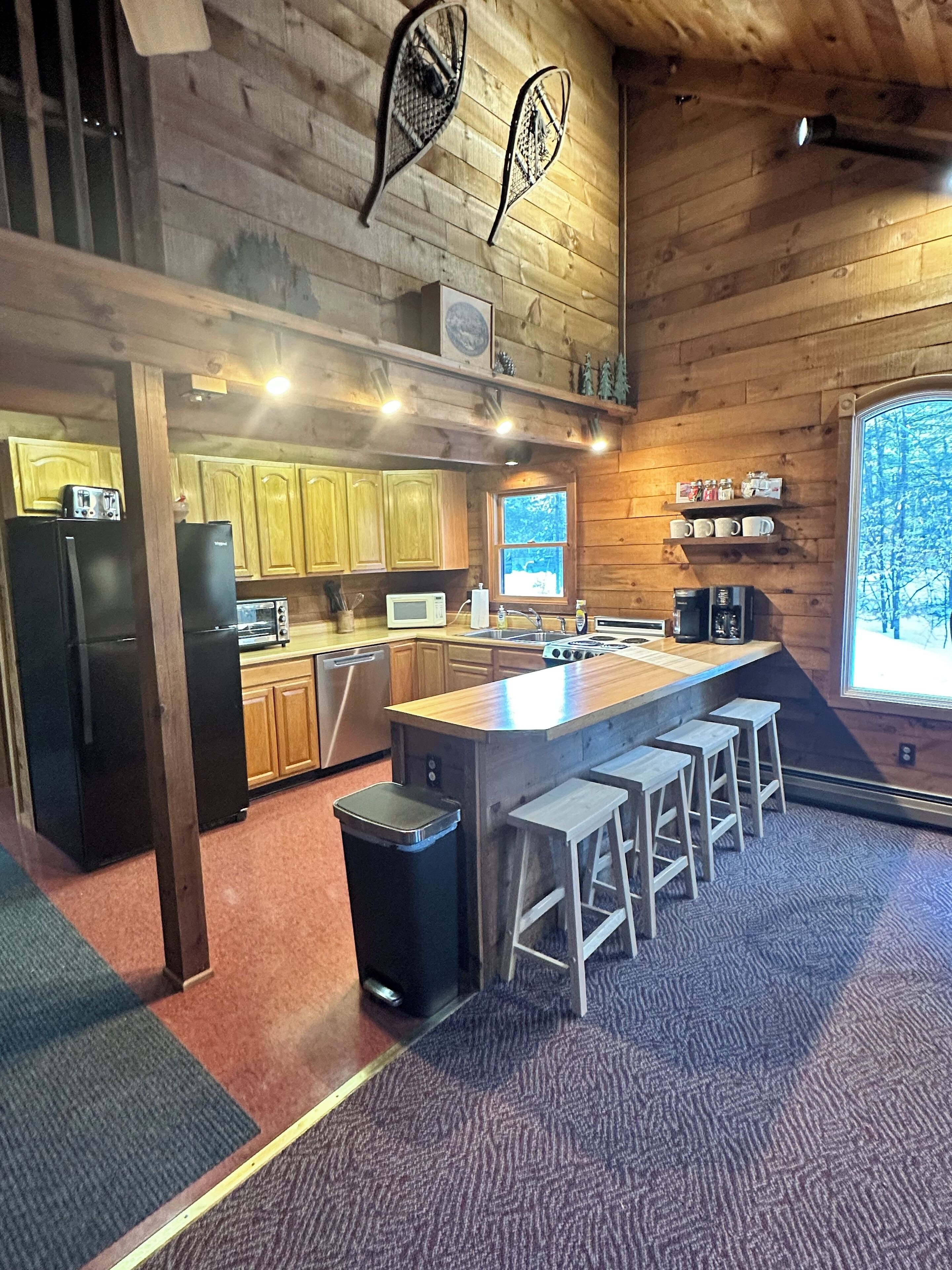 A modern kitchen with wooden cabinets, a central island with stools, a refrigerator, a microwave, and a coffee machine.