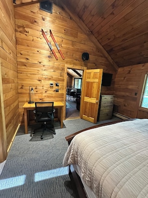 Un dormitorio acogedor en el ático con paredes de madera, un escritorio con silla, una cama y una puerta que da a otra habitación.