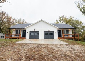 A two-story building with a central garage entrance, wooden balconies, and a concrete driveway. - BRAND NEW 3BR Duplex Unit Side “B”, Retreat Near Fort Leonard Wood,Sleeps 8 (Waynesville)