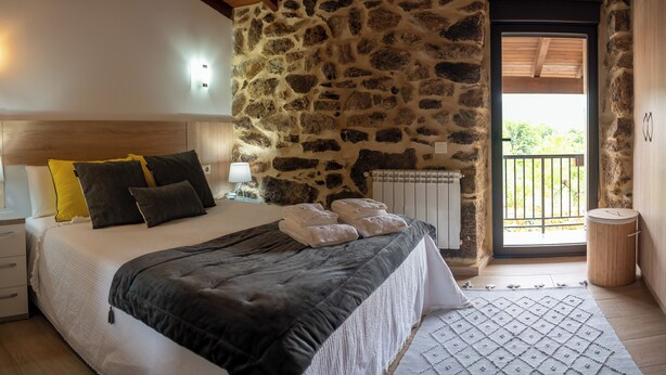 A bedroom with a stone wall, a bed with a dark blanket, a wooden headboard, and a view of the outdoors. - Complete 4 Bedroom House near Termas de Bande and Lobios - Villa Limia (Chaos)