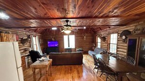 A rustic cabin interior with a wooden ceiling, a ceiling fan, a television, a sofa, a dining table with chairs, and a refrigerator.