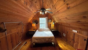 A cozy attic bedroom with wooden walls, a bed, two bedside tables with lamps, a ceiling fan, and a window.