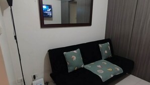 Ein Zimmer mit einer schwarzen Couch, zwei gemusterten Kissen, einer stehenden Stehlampe und einem an der Wand montierten Fernseher.